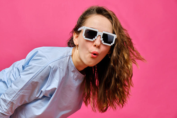 Stilvolles kaukasisches Mädchen in blassblauem T-Shirt und 8-Bit-Brille und macht einen überraschten Blick auf rosa Hintergrund. Nahaufnahme. - Foto, Bild