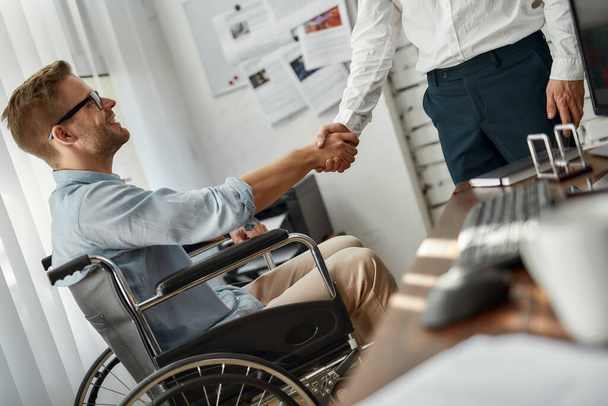 Welcome to our team. Cropped view of young male office worker in a wheelchair shaking hand to his colleague while working together in the creative office. Disability concept - Фото, изображение