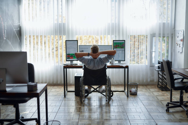 Back view of relaxed young male trader in a wheelchair keeping hands behind head and looking at three computer monitors with diagrams and graphs while working in modern office - Fotoğraf, Görsel