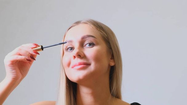Woman puts black mascara on her eyelashes with makeup, photo of attractive blonde on a white and gray background. - Foto, Imagem