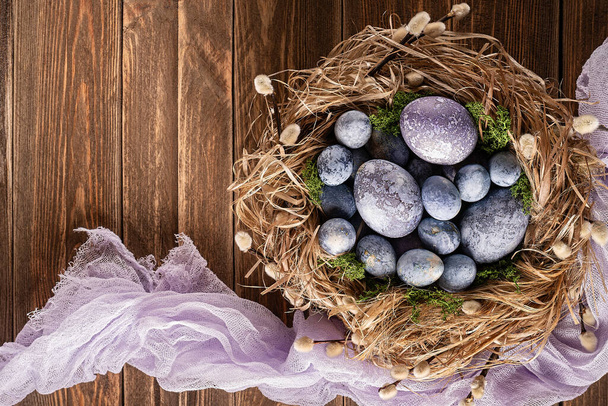 Colorful easter eggs in a nest with a light purple cheesecloth on a wooden background. Easter card. Top view. Copy space.  - Photo, Image