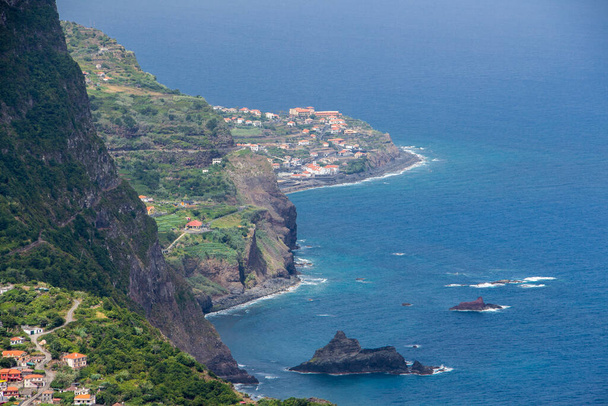 Kayalıklardan Portekiz, Madeira 'nın kuzey kıyısındaki küçük Ponta Delgada şehrine kadar. Adanın bir burnu üzerinde uzanmış denizi işaret eden, etrafını saran yüksek dağlarla tipik medreseli köyler ve evler.. - Fotoğraf, Görsel