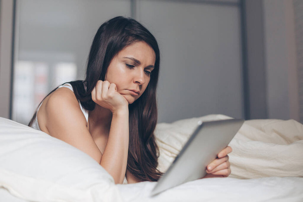 Sad girl looks into tablet in hands on bed - Valokuva, kuva