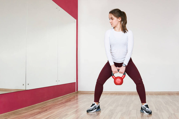 young petite woman is training with kettlebell in a spacious gym looking in the mirror - Photo, Image