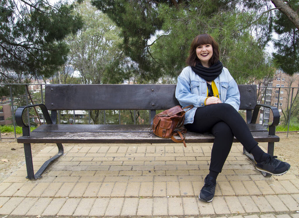 明るい現代的な服のベンチに座っている都市の若いスタイリッシュな笑顔の女性の肖像画。ライフスタイルと都市概念. - 写真・画像