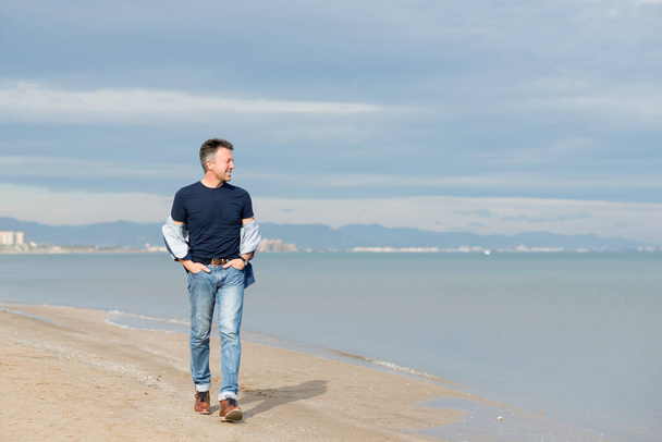 Handsome middle-aged man walking at the beach. Attractive happy smiling mid adult male model posing at seaside in blue jeans, t-shirt shirt. Full-length outdoor portrait of beautiful macho man.  - Фото, изображение