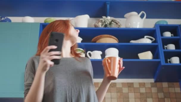 Dancing fun, listening to music on smartphone. Attractive girl with red hair drinks morning coffee in the home kitchen. Young female dancing and using cellphone. Redhead woman dancing in the kitchen - Filmagem, Vídeo