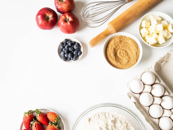 Frame of various baking ingredients - flour, eggs, sugar, butter, fresh berries, nuts, kitchen utensils on white background. Top view. Copy space - Photo, Image