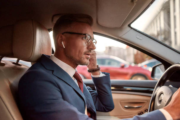 Hablando con el cliente. Hombre de negocios guapo en traje completo ajustando auriculares mientras conduce un coche
 - Foto, Imagen