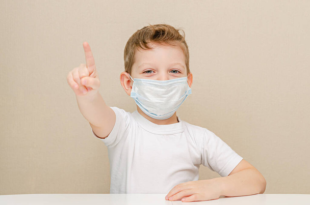 Netter Junge von vier Jahren in einer medizinischen Maske. Kinder aufgrund einer Epidemie unter Quarantäne gestellt. Der Junge bekam eine Idee. Geste - Daumen hoch, Aufmerksamkeit - Foto, Bild