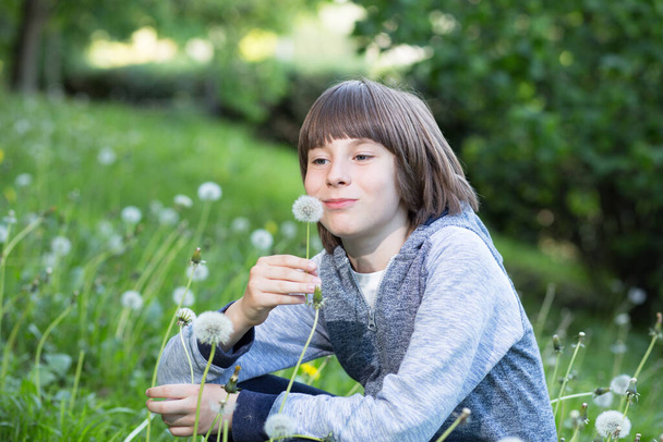 Мальчик дует одуванчик над размытой зеленой травой, летняя природа на свежем воздухе. Детство, мечты, лето, хорошее настроение
. - Фото, изображение