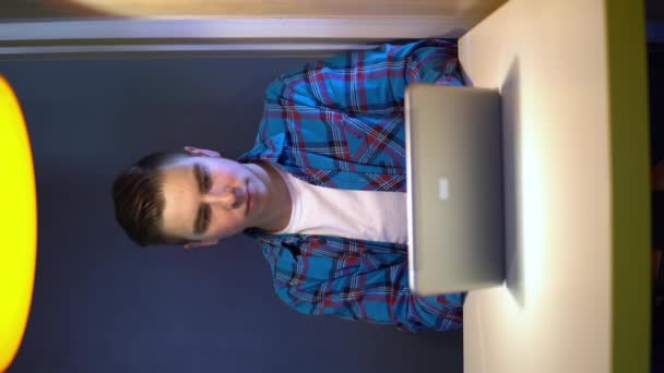 Nuori mies kannettavan kanssa istuu pöydässä. Mies istuu kahvilassa pöydässä pienen kannettavan tietokoneen kanssa ja kirjoittaa kirjeitä. Pystysuora video
. - Materiaali, video