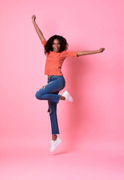 Radosna młoda afrykańska kobieta w pomarańczowej koszuli skacze i świętuje odizolowane na ponad różowym tle studio. - Zdjęcie, obraz