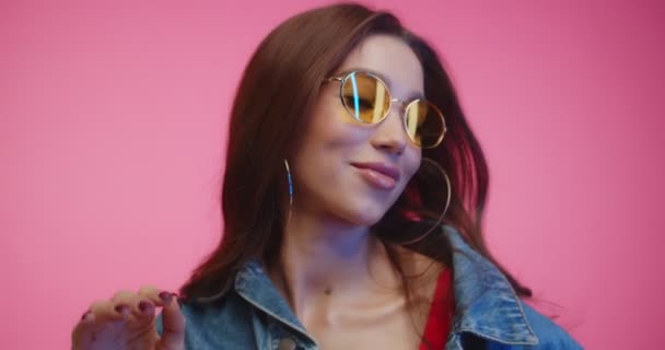 Sassy girl in yellow glasses looking to camera and dancing. Portrait of fashion girl dancing in pink studio. Close up of young woman in sunglasses looking and smilingw hile dancing on pink background - 映像、動画