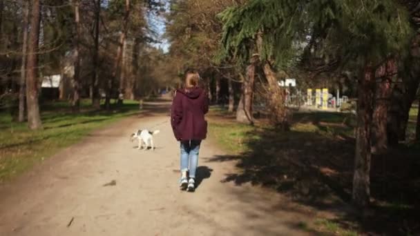 Beautiful girl with a backpack on her back walks her dog on a leash in the spring park. Dog walk, village life, pet care, happy weekend - Materiał filmowy, wideo
