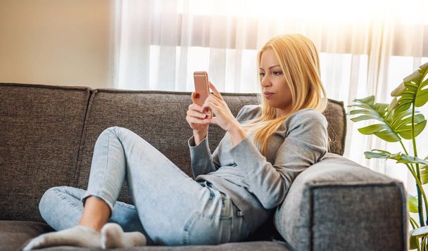 自宅でソファに座っている間に携帯電話を使う女性リビングルームでスマートフォンを使うかなりの女性. - 写真・画像