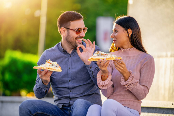 Φωτογραφία του ευτυχισμένου νεαρού ζευγαριού τρώει πίτσα σε εξωτερικούς χώρους.Fast food έννοια. - Φωτογραφία, εικόνα