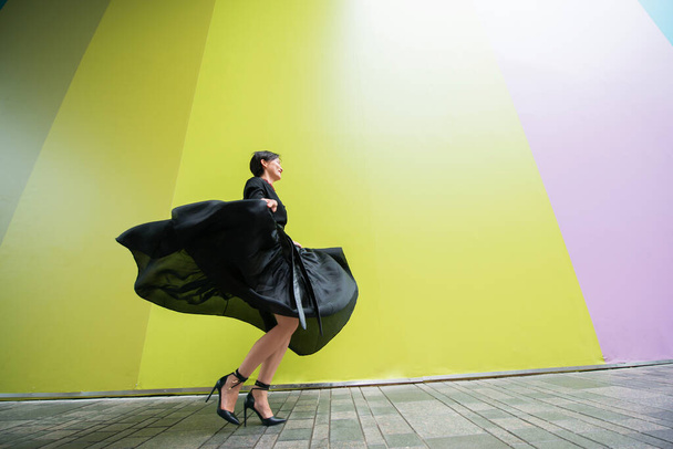 Femme de mode. Jeune belle fille chinoise dansant en plein air portant une longue robe noire avec des talons hauts sur fond de mur coloré. Dame à la mode élégant.  - Photo, image