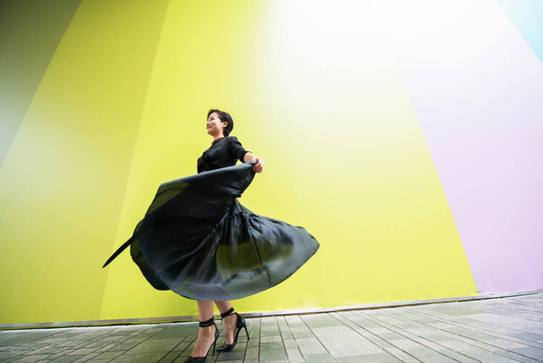 Fashion woman. Young beautiful chinese girl dancing outdoor wearing long black dress with high heels over colorful wall background. Stylish trendy lady.  - Φωτογραφία, εικόνα
