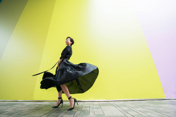 Fashion woman. Young beautiful chinese girl dancing outdoor wearing long black dress with high heels over colorful wall background. Stylish trendy lady.  - Zdjęcie, obraz