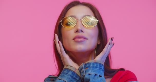 Sassy model in yellow glasses looking to camera and dancing. Face potrait of fashion girl in pink studio. Close up of young woman in sunglasses looking and smiling to camera on pink background. - Filmati, video