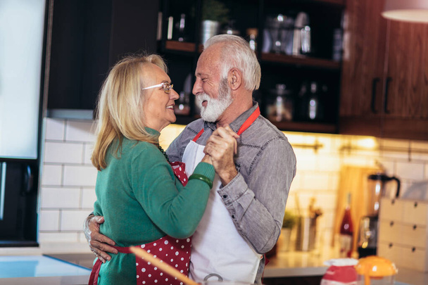 Beautiful senior couple is dancing and smiling while cooking together in kitchen. Quarantine. Health concept. - Photo, Image