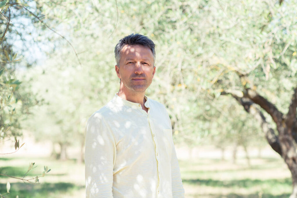 Olivos. Hombre guapo posando en el jardín de olivos. Masculino portrain sobre campo de olivo mediterráneo listo para la cosecha. Hombre maduro y seguro en el olivar italiano con aceitunas frescas maduras. Aceitunas frescas. Explotación oleícola
 - Foto, imagen