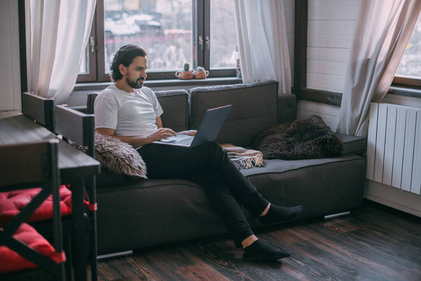 Kotona töissä. Mies työskentelee kannettavalla tietokoneella kotona, istuu olohuoneen sohvalla päivän aikana, puhuu puhelimessa. Työskentele kotoa
. - Valokuva, kuva