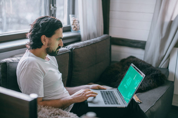 Thuis werken. Een man werkt thuis op een laptop, zit overdag op een bank in de woonkamer, praat aan de telefoon. Werk vanuit huis. - Foto, afbeelding