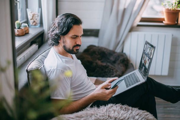 Arbeitsplatz zu Hause. Ein Mann arbeitet zu Hause am Laptop, sitzt tagsüber auf einem Sofa im Wohnzimmer, telefoniert. Arbeit von zu Hause aus. - Foto, Bild