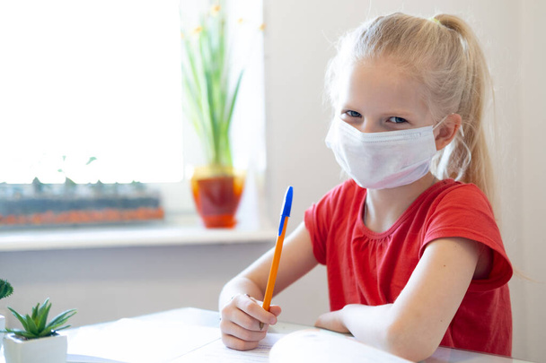 Serios sad blonde schoolgirl in medical mask studying at home, doing school homework, writing in notebook. Reading training books on table. coronavirus quarantine. Distance learning online education. - Photo, Image