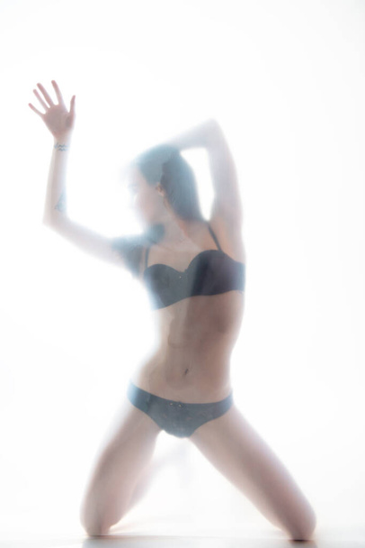 Young beautiful woman posing in lingerie behind translucent material. Full-length attractive sexy girl's portrait in studio over white background, foggy silhouette. Art female portrait. - Foto, afbeelding