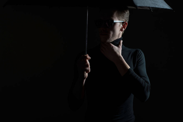 Dramatic studio portrait of a guy in black, holding an umbrella in sunglasses. On a gray background. Hide from the rain under an umbrella. Advertising umbrella. Backlight Guy in black clothes. Portrait of a dark man - Foto, afbeelding