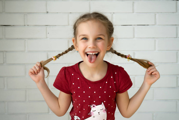 Adorable chica divertida sosteniendo coletas de pelo en una pared de ladrillo. Pequeña linda chica con el pelo largo y rubio al aire libre jugando, mostrando la lengua
 - Foto, Imagen