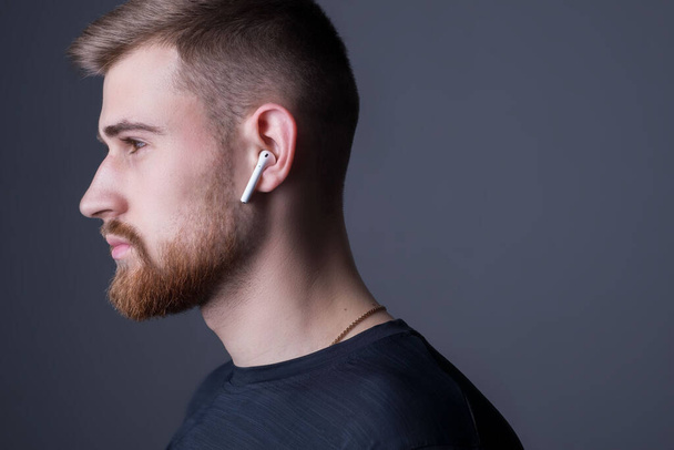 Closeup portrait of a young bearded guy of twenty-five years old, standing in profile. In wireless white headphones. Sporty style. Sutdiy advertising photo of the athlete in the headphones. Sports and music. Guy listens to music on headphones - Foto, afbeelding