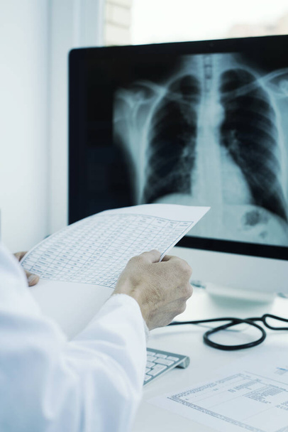 Крупный план молодого врача в белом халате, сидящего за столом, проверяющего результаты теста и рентгенограммы грудной клетки в компьютере
 - Фото, изображение