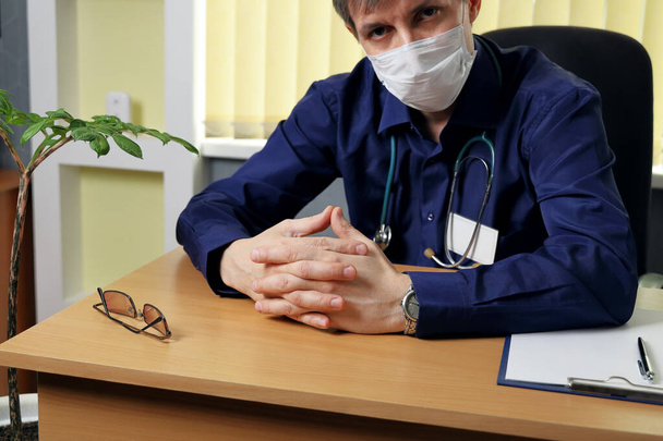 Ένας γιατρός με ιατρική μάσκα κάθεται σε ένα τραπέζι. Ακούει προσεκτικά τον συνομιλητή. - Φωτογραφία, εικόνα