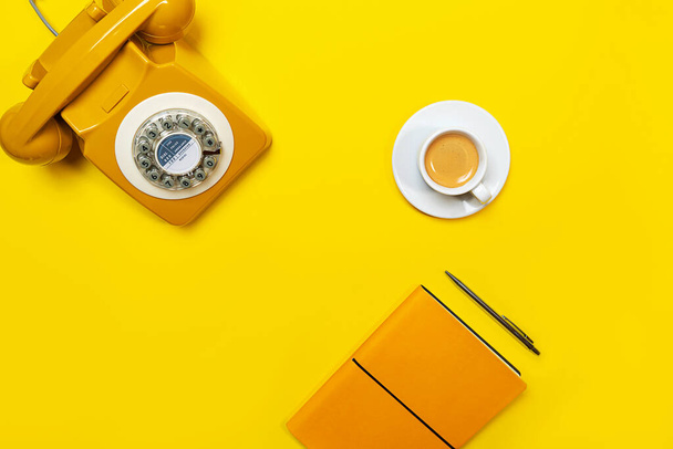 Yellow layout with retro rotary telephone, cup of coffee, pen and notebook. - 写真・画像