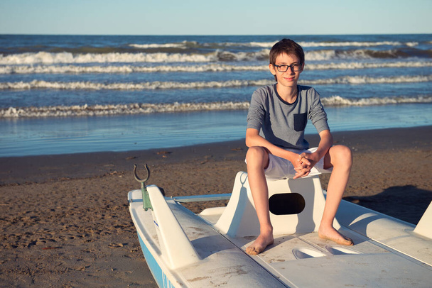 Young boy sitting on catamaran at the summer beach. Cute spectacled smiling happy 12 years old boy at seaside, looking at camera. Kid's outdoor portrait over seaside. - 写真・画像