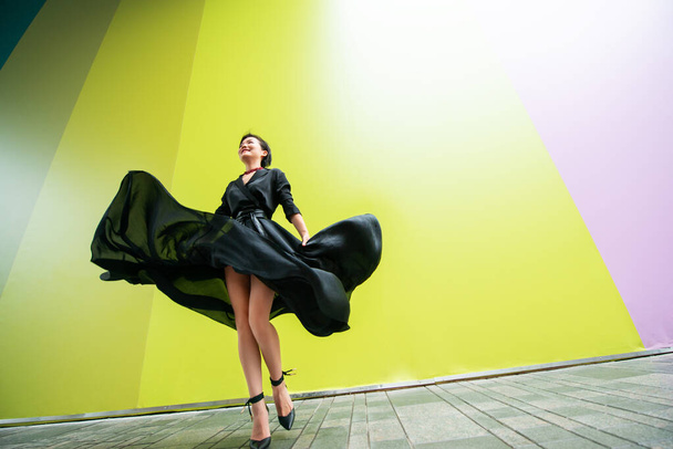 Fashion woman. Young beautiful chinese girl dancing outdoor wearing long black dress with high heels over colorful wall background. Stylish trendy lady.  - 写真・画像