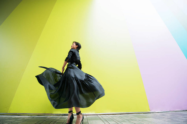 Fashion woman. Young beautiful chinese girl dancing outdoor wearing long black dress with high heels over colorful wall background. Stylish trendy lady.  - Foto, imagen