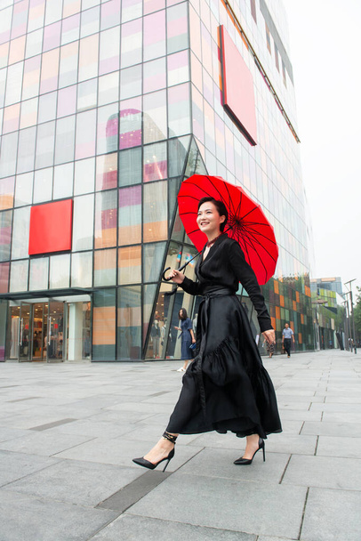 Fashion woman walking in center of Beijing. Young beautiful chinese girl posing outdoor wearing long black dress with high heels holding red umbrella over colorful wall background. Stylish trendy lady. - Fotó, kép