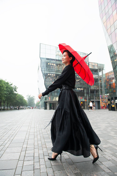 Fashion woman walking in center of Beijing. Young beautiful chinese girl posing outdoor wearing long black dress with high heels holding red umbrella over colorful wall background. Stylish trendy lady. - Photo, Image