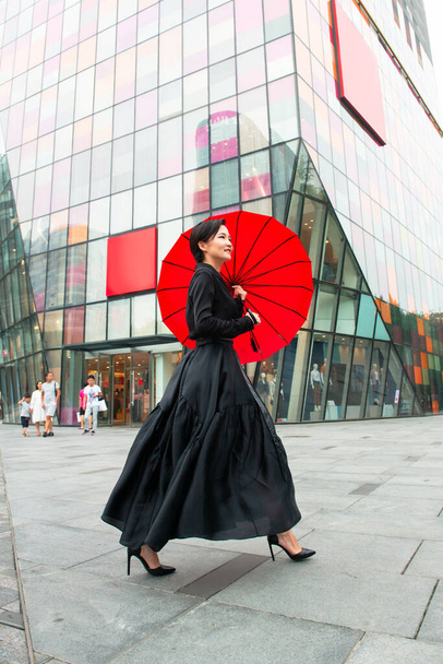 Fashion woman walking in center of Beijing. Young beautiful chinese girl posing outdoor wearing long black dress with high heels holding red umbrella over colorful wall background. Stylish trendy lady. - Фото, зображення