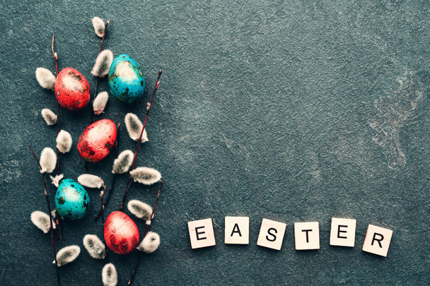 Bright red and blue easter quail eggs and fluffy willow branches, text EASTER made from wooden letters on a black background, top view. Easter background. - Photo, Image
