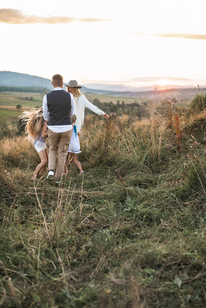 Mom, dad and little two daughters have fun in summer field together at sunset. They run, hug each other and have fun together. Happy family - Zdjęcie, obraz
