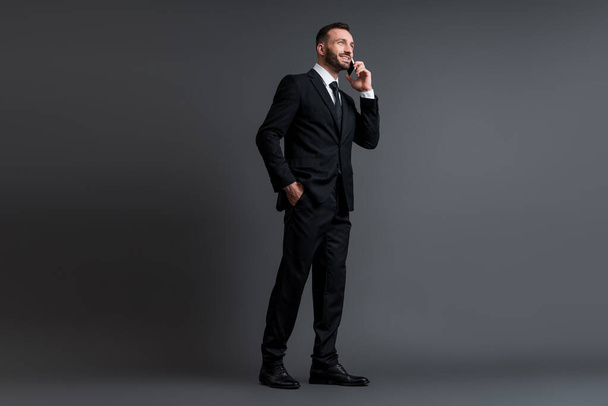 happy businessman in suit talking on smartphone while standing with hand in pocket on grey  - Fotografie, Obrázek