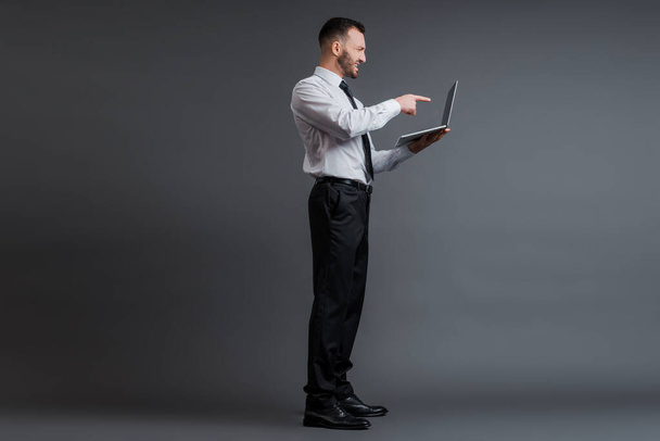 προφίλ θυμωμένου επιχειρηματία με κοστούμι που δείχνει με το δάχτυλο το φορητό υπολογιστή σε γκρι  - Φωτογραφία, εικόνα