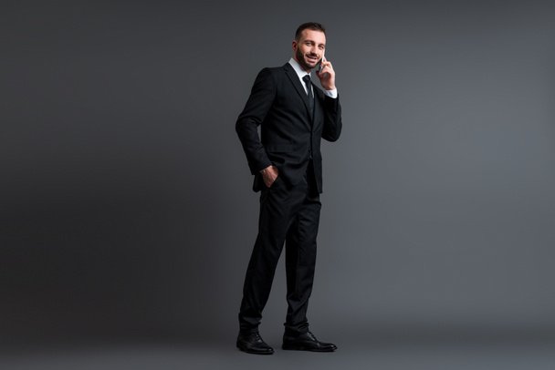 cheerful businessman in suit talking on smartphone while standing with hand in pocket on grey  - Zdjęcie, obraz