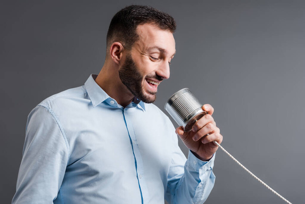 happy bearded man talking while holding tin can isolated on grey  - Zdjęcie, obraz